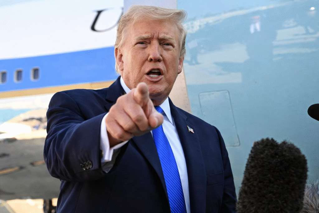 Donald Trump speaking outdoors near a large aircraft, gesturing with his hand while addressing reporters, with a microphone visible in the foreground.