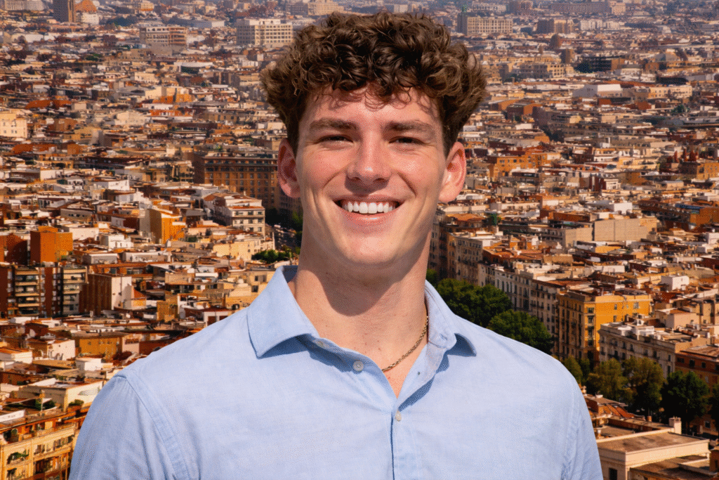 Young man with curly brown hair smiling in front of a dense Barcelona cityscape during daylight