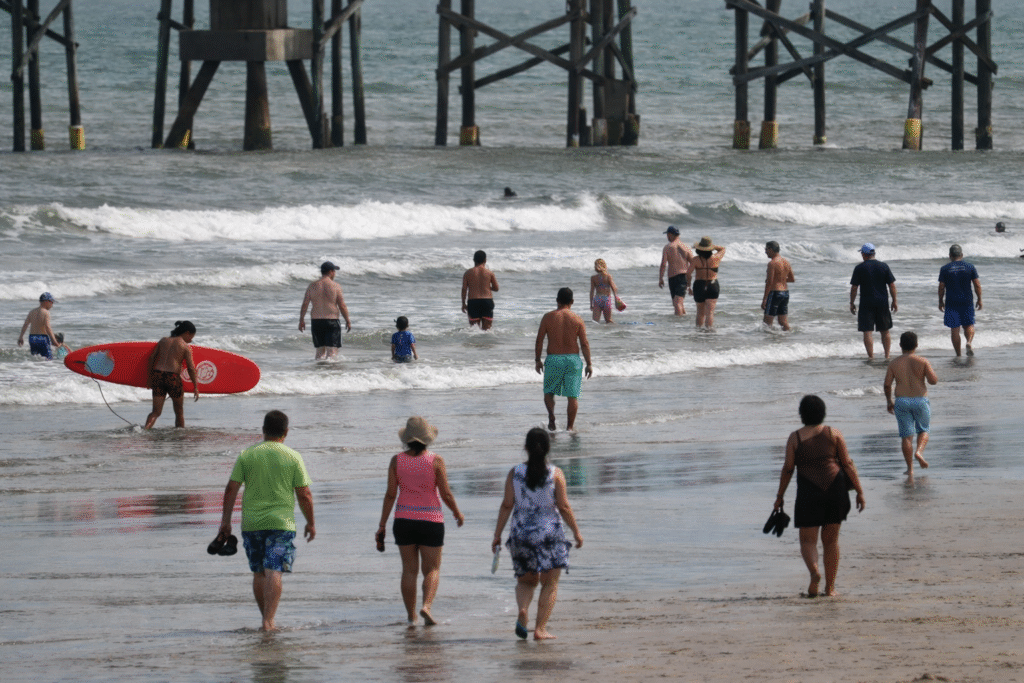 People walking and wading in shallow ocean water along a sandy beach during daytime, with small waves and a wooden pier structure visible in the background.