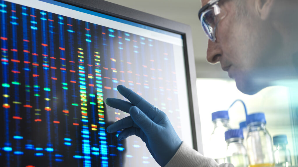 Scientist wearing gloves examining a computer display showing colored DNA sequencing data in a biotechnology laboratory.