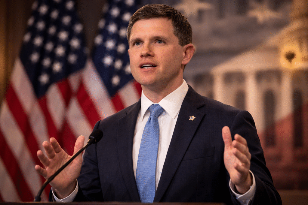 Texas Democrat James Talarico speaking at a press conference podium with American flags in the background, gesturing with his hands during a political statement.