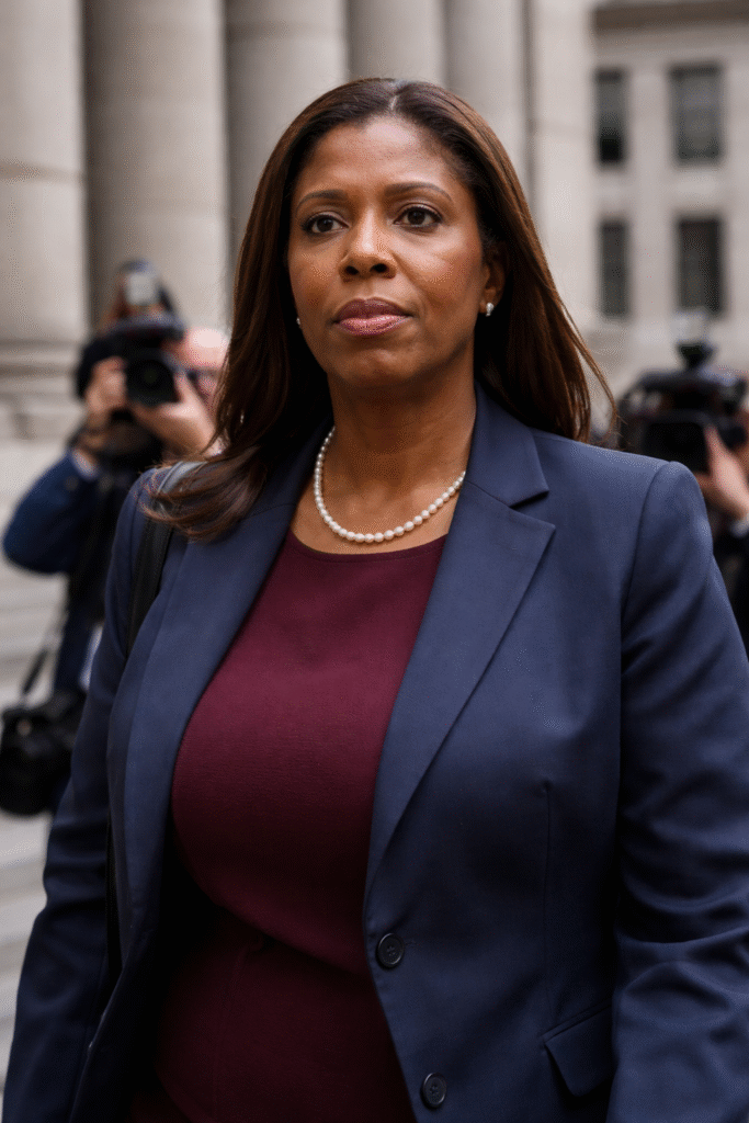 Close-up of Letitia James walking outside a courthouse, wearing a navy blazer and burgundy top, with blurred reporters and cameras in the background.