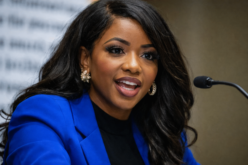 Female politician speaking at a microphone during a public event, wearing a blue blazer and addressing an audience at a press conference.