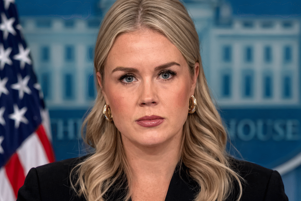 Karoline Leavitt speaking in the White House press briefing room with the White House emblem and an American flag visible behind her.