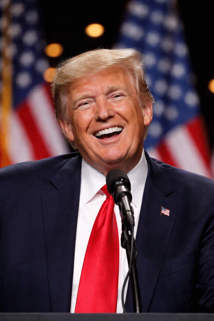 President Donald Trump smiling and laughing at a podium during a public event, with American flags in the background and stage lighting behind him