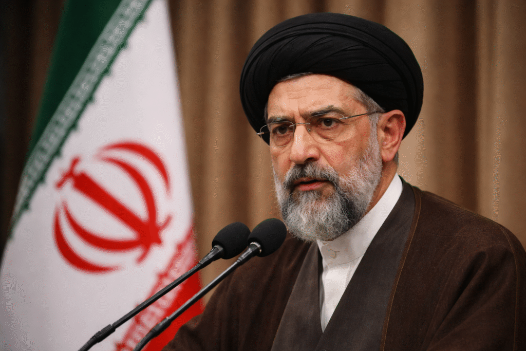 Mojtaba Khamenei speaking at a podium wearing a black turban and clerical robe with the Iranian flag visible in the background.