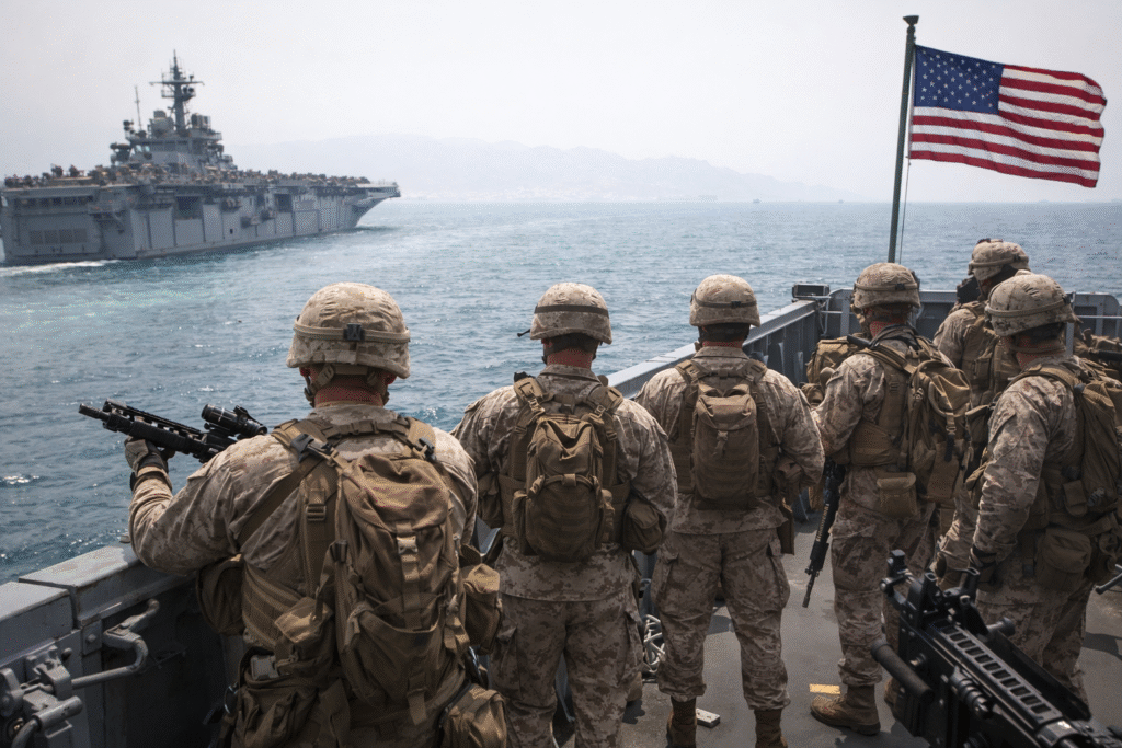 U.S. Marines in combat gear standing on a naval ship deck facing the sea, with a large warship and American flag visible in the background