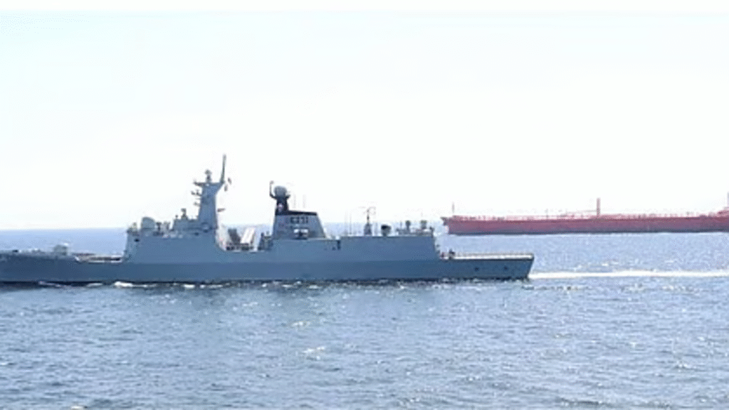 U.S. Navy destroyer sailing across open water with a large commercial oil tanker visible in the background along a major maritime shipping route.