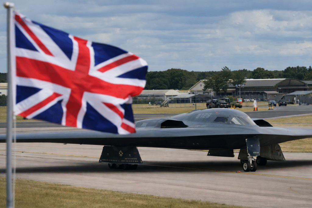 U.S. B-2 Spirit stealth bomber parked on a runway at a British RAF airbase with a Union Jack flag in the foreground.