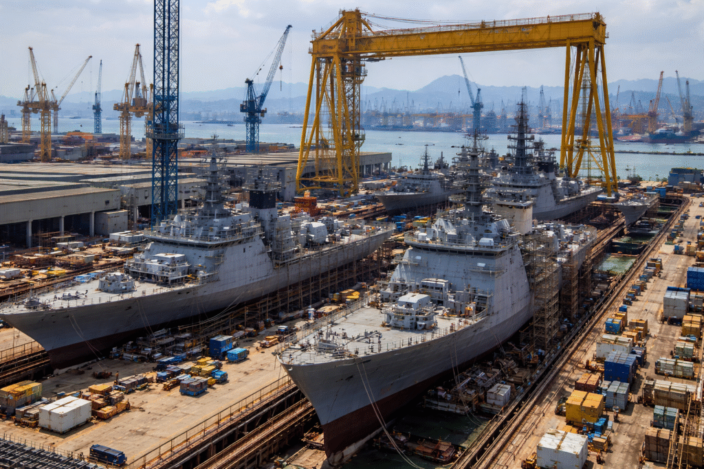 Large industrial shipyard with multiple naval warships under construction in dry docks, surrounded by heavy cranes and manufacturing infrastructure.
