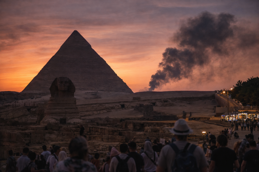 Tourists walking near the Great Sphinx and Pyramid of Giza at sunset, with market lights and a plume of smoke visible in the background.