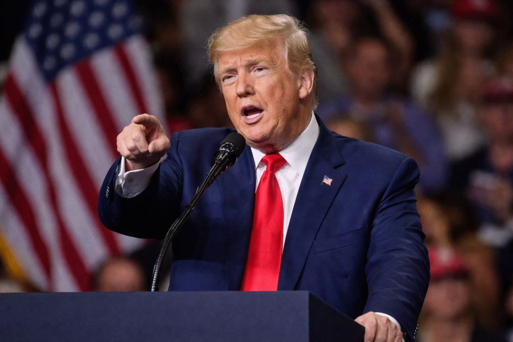 President Donald Trump speaking at a political rally, pointing while standing behind a podium with American flags and a crowd blurred in the background.