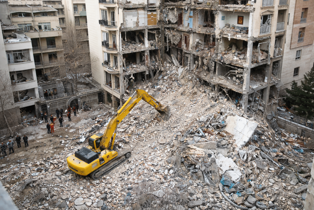 Damaged multi-story residential building with exposed interiors and large rubble pile, as a yellow excavator clears debris while rescue workers stand nearby in an urban area
