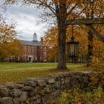 Quiet college campus in autumn with empty lawn, trees with orange leaves, and a distant academic building symbolizing institutional decline