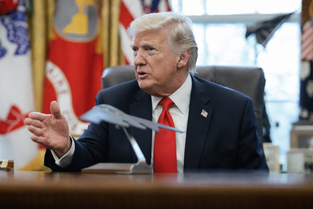 A man in a dark suit with a red tie sits at a desk, gesturing with one hand while speaking, with flags and a model aircraft visible in the background.