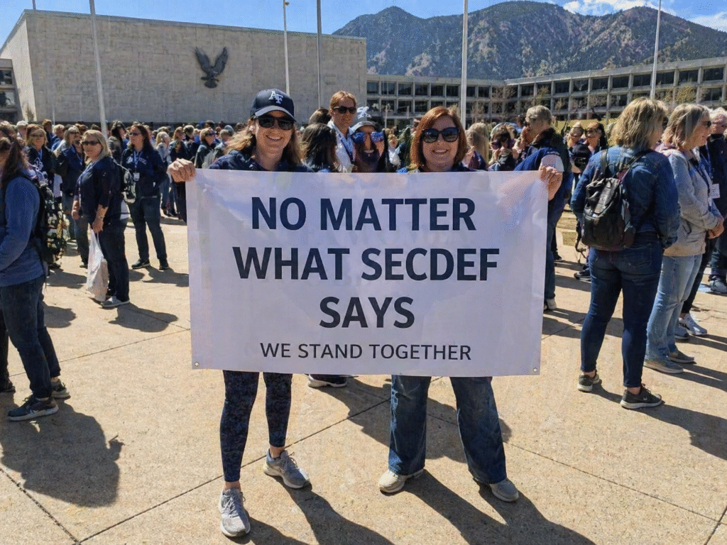 Group of women gathered outside a large institutional building holding a banner reading “No matter what SecDef says we stand together,” with a crowd and mountain backdrop in daylight.