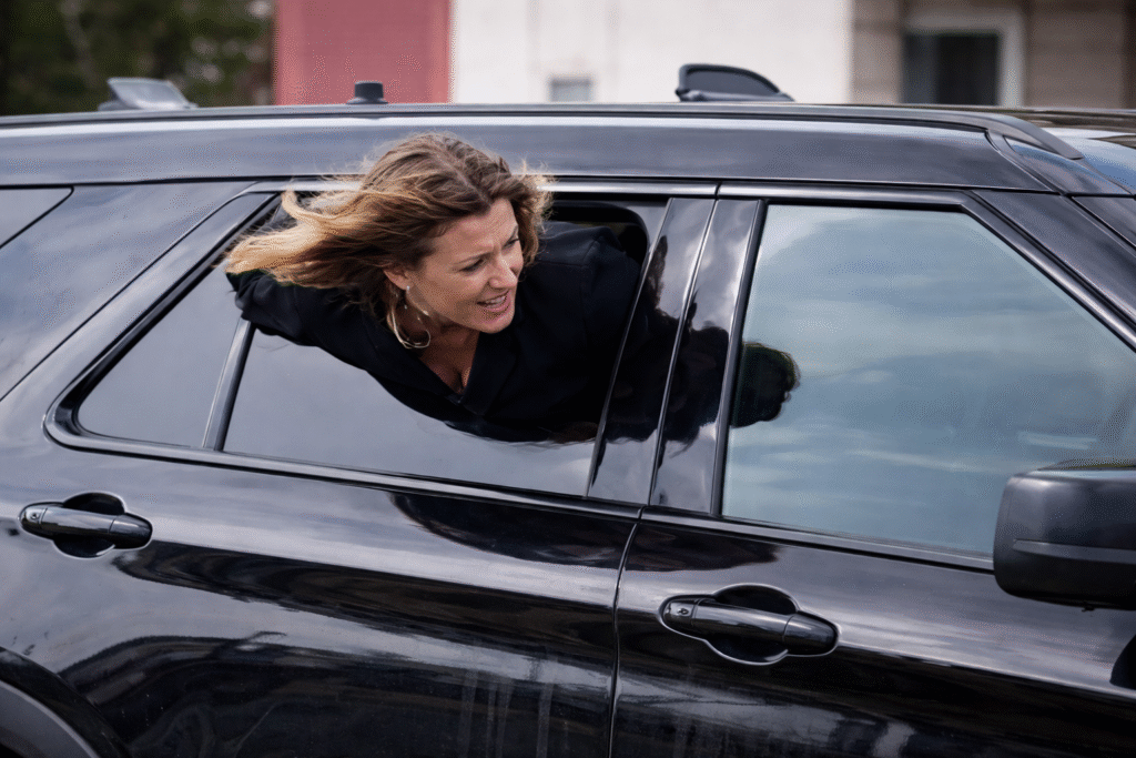 Woman leaning out of a partially open rear window of a dark SUV, upper body outside the vehicle while attempting to exit, with an outdoor parking area in the background.