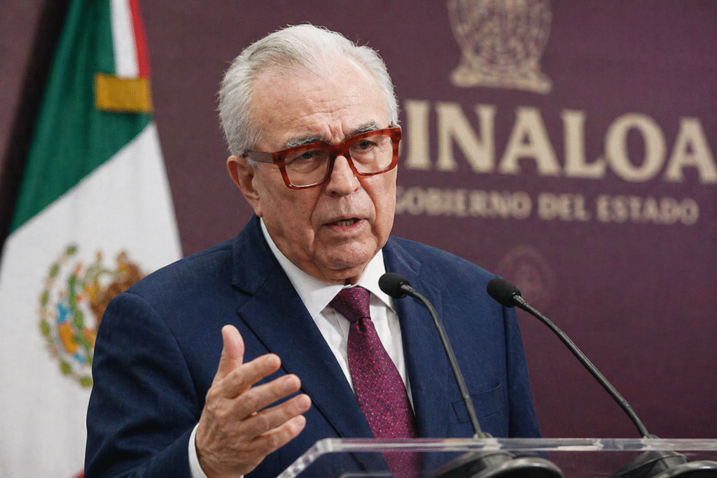 Older man in a suit and glasses speaking at a podium during a press briefing, gesturing with his hand against a blurred official backdrop.