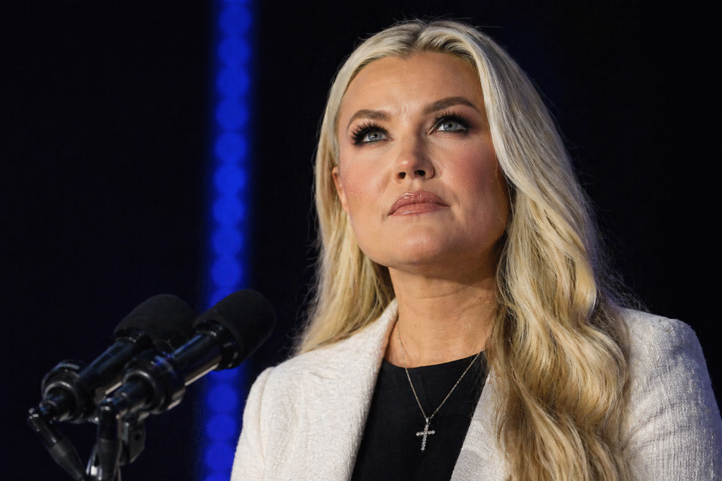 Blonde woman standing at a podium with microphones, looking upward under stage lighting in a dark indoor setting.