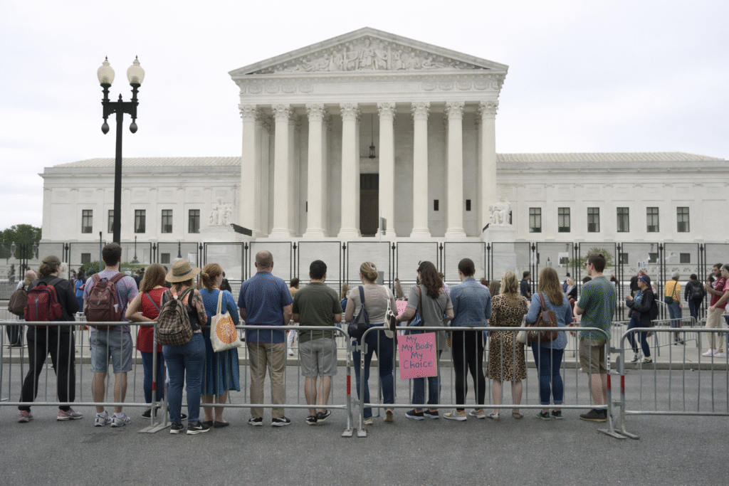 Crowd of people standing behind metal barricades outside the US Supreme Court building under an overcast sky, with varied clothing and postures, viewed from behind.