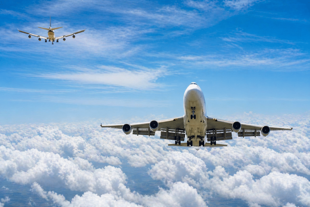 Two commercial passenger airplanes flying at different altitudes above a dense layer of clouds, with one large jet in the foreground and another approaching from the distance under a clear blue sky.