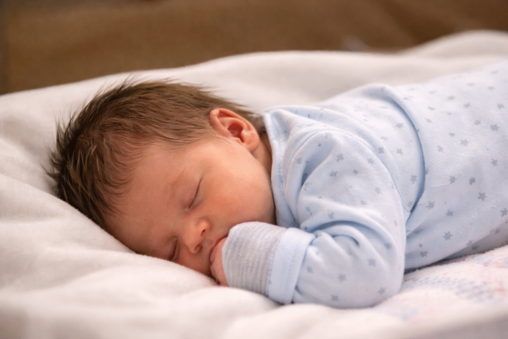 Sleeping newborn baby lying on a soft blanket, wearing a light blue onesie, resting peacefully on their side with one hand near their face.