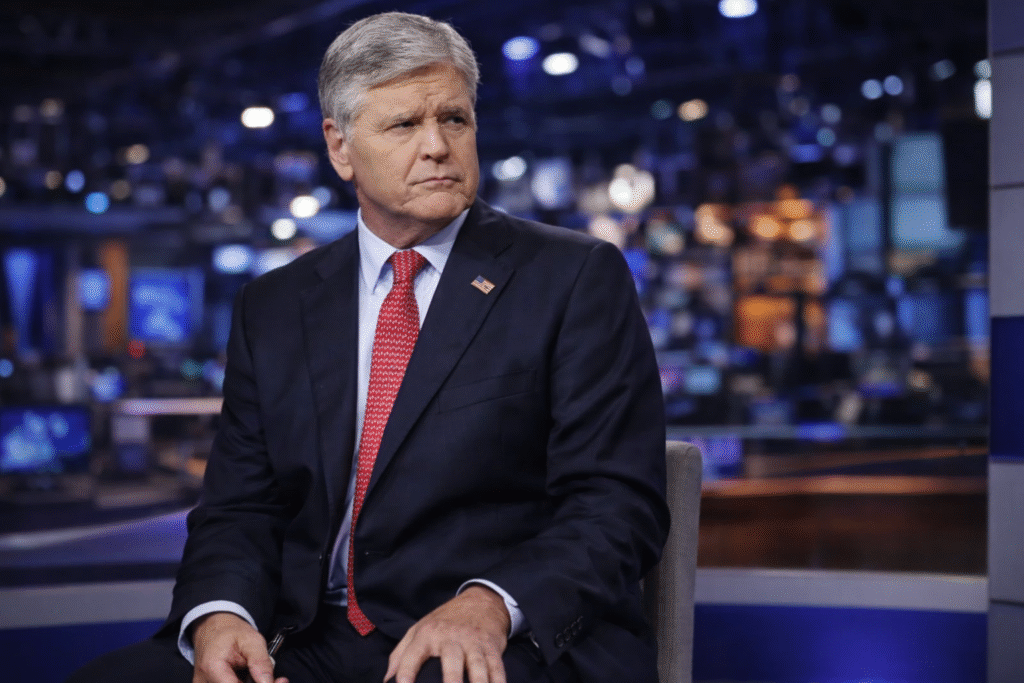 Professional news studio setting with a middle-aged male presenter in a navy suit and red tie, seated and looking to the side with a serious expression, against a softly blurred background of studio lights and screens.