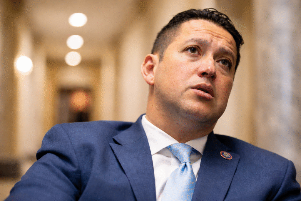 Close-up of a man in a navy suit and light blue tie seated indoors, with a blurred government-style hallway in the background and a serious expression on his face.
