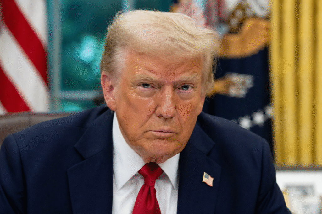 Portrait of Donald Trump seated indoors in a dark suit and red tie, with a blurred American flag and office setting in the background.