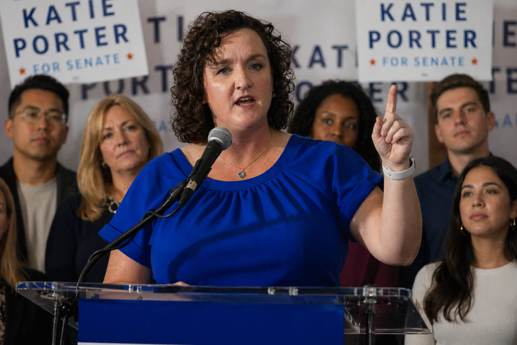 Woman speaking at a podium during a campaign event, raising her finger while addressing an audience, with supporters standing behind her holding signs.