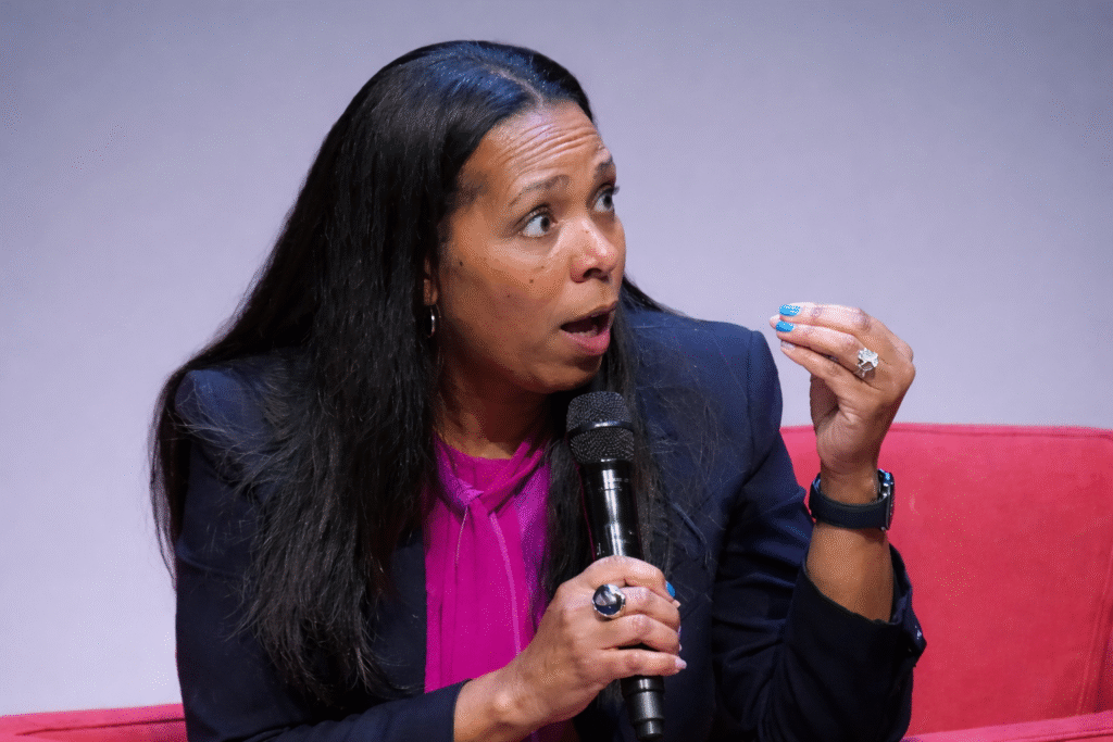 Woman speaking into a handheld microphone during a panel discussion, wearing a navy blazer and magenta blouse against a neutral gray background, gesturing while addressing an audience.