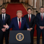 Man at a presidential podium gesturing with one hand while speaking, with three suited officials standing behind him in a formal government hallway lined with columns and a red carpet.