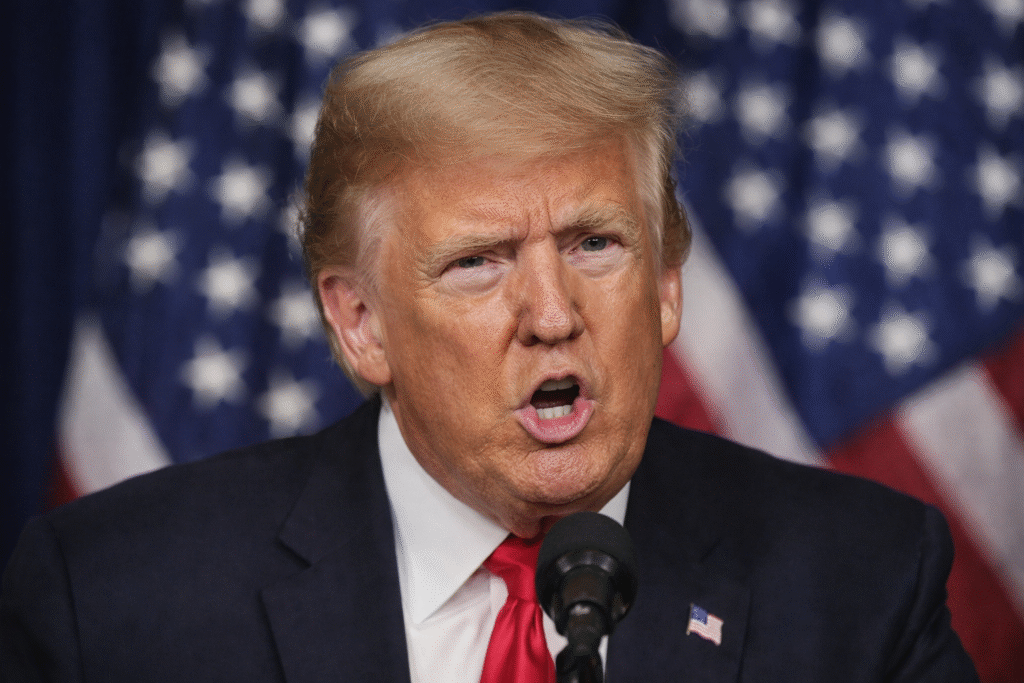 Donald Trump speaking at a podium with an American flag backdrop, wearing a dark suit and red tie, captured mid-speech.