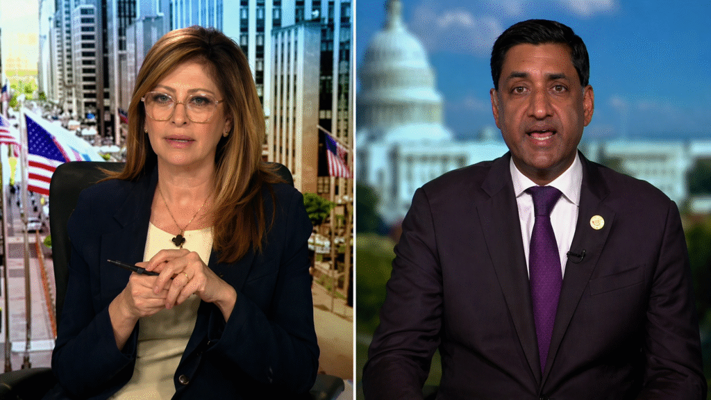 Split-screen television news interview showing a female anchor in a studio on the left and a male guest speaking from a remote location with a blurred government building in the background.