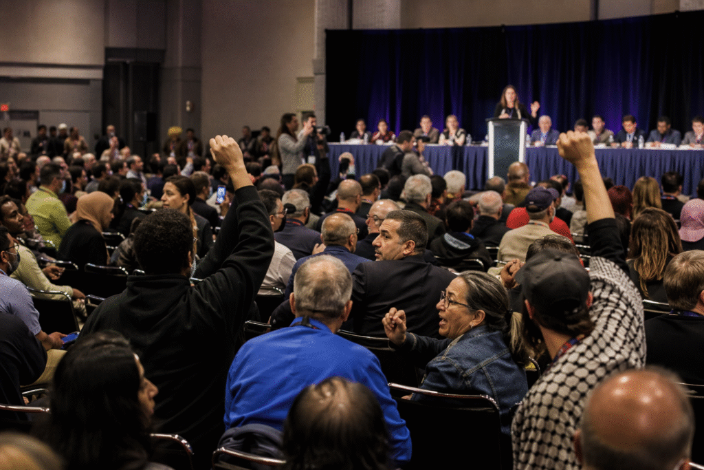 Crowded political convention hall with a speaker at a podium and delegates reacting with visible disagreement and tension.