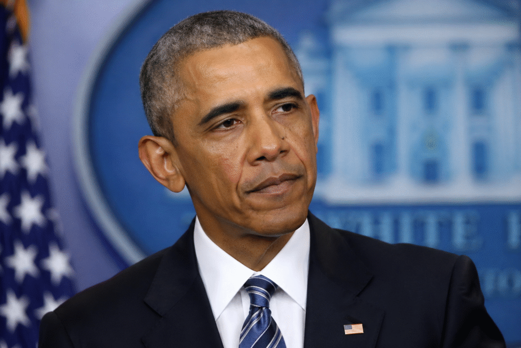 Portrait of Barack Obama in a dark suit and striped tie, photographed indoors against a blurred blue background with a faint U.S. flag visible.