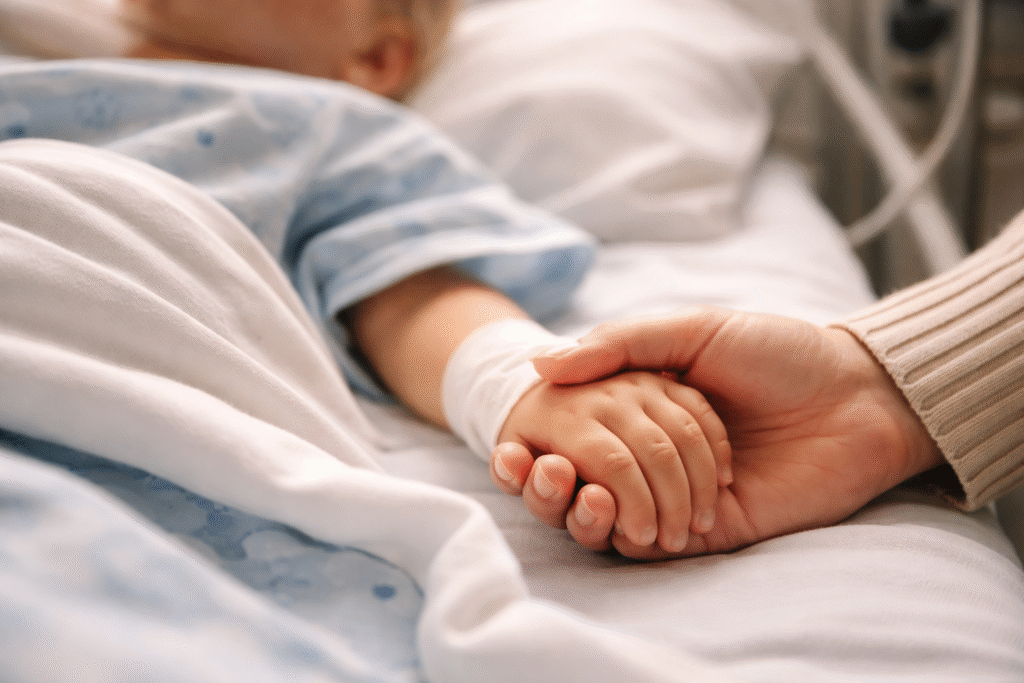 Close-up of an adult gently holding a toddler’s hand in a hospital bed, with the child’s face out of focus, conveying care and recovery.