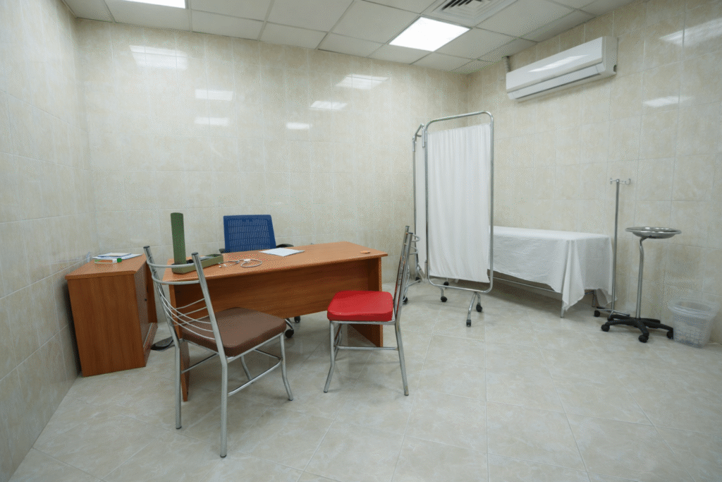 Empty medical consultation room with beige tiled walls, desk with chairs, examination bed, privacy screen, and clinical equipment in a clean, sterile environment.