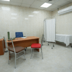 Empty medical consultation room with beige tiled walls, desk with chairs, examination bed, privacy screen, and clinical equipment in a clean, sterile environment.