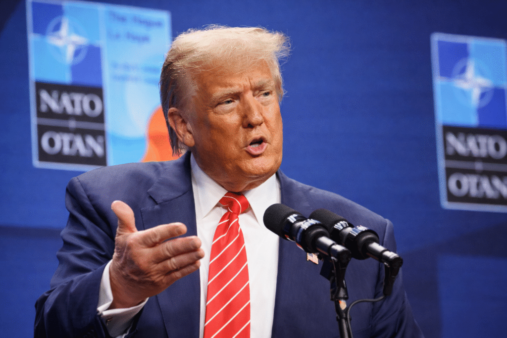 Man in a navy suit and red striped tie speaking at microphones during a NATO event, gesturing with one hand against a backdrop of NATO logos.