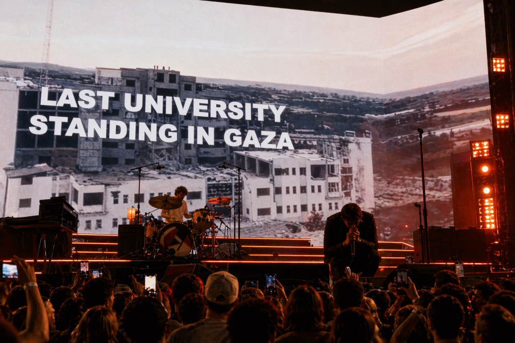Wide shot of a live concert stage with a band performing under warm orange lighting, while a large LED screen behind them displays a damaged building and bold text referencing a university in Gaza, with a crowd in the foreground holding up phones.