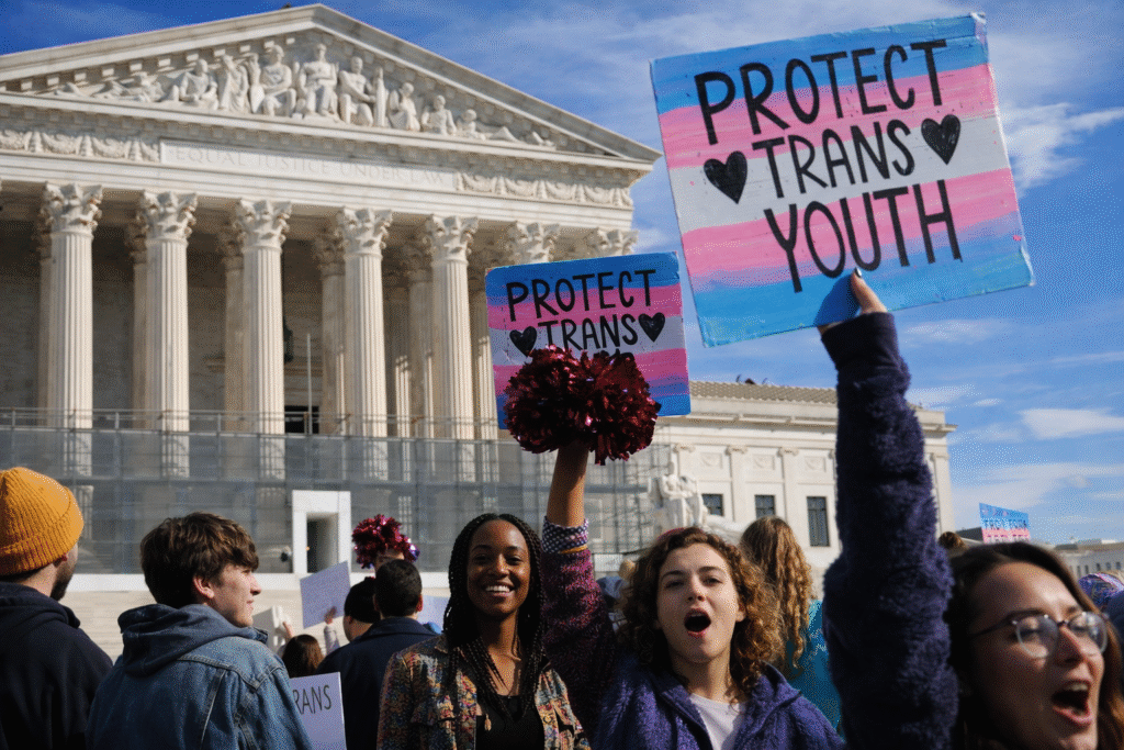 Group of protesters gathered outside the U.S. Supreme Court building holding colorful signs supporting transgender youth, with raised arms and a crowd facing the courthouse under a clear sky.