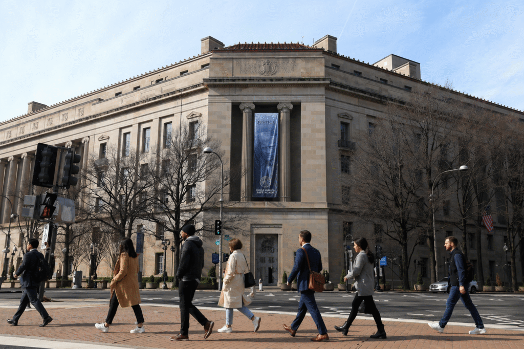 Street-level view of a large neoclassical government building with columns as a group of pedestrians in varied coats and outfits walk across a crosswalk in front.