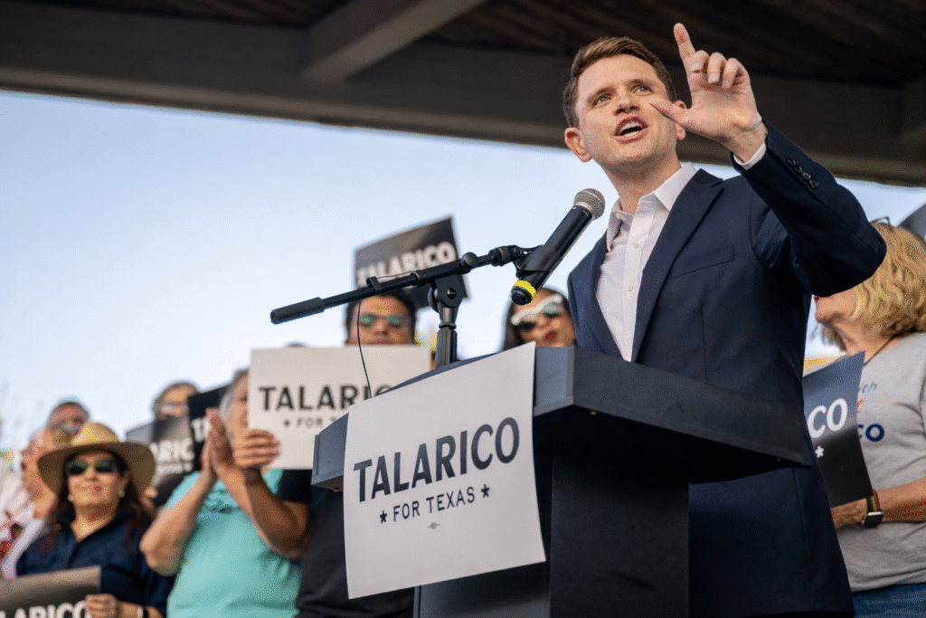 A man in a navy suit speaks at an outdoor campaign rally from behind a podium, gesturing as supporters hold signs in the background.