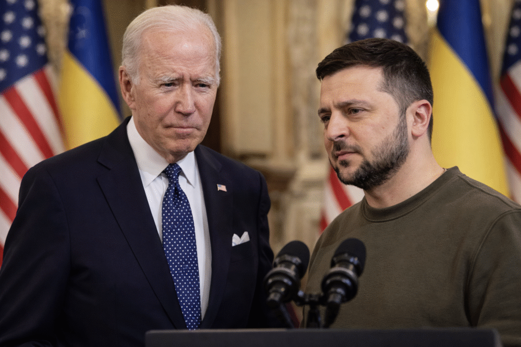 Joe Biden and Volodymyr Zelensky stand side by side at a podium with microphones, with U.S. and Ukrainian flags in the background during a formal event.