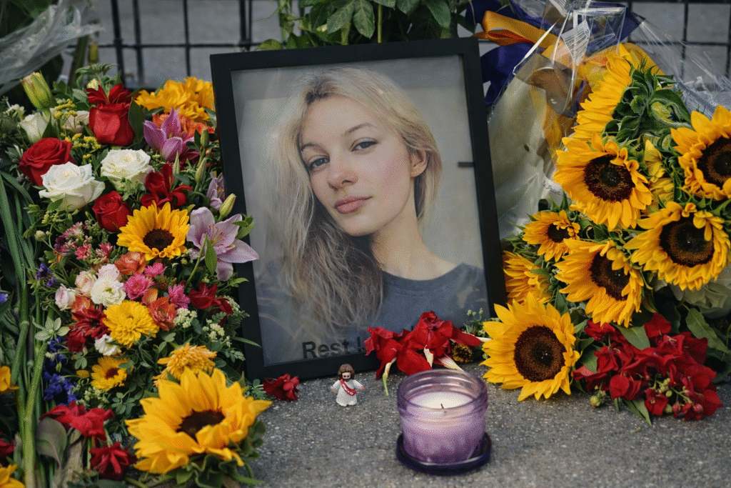 Memorial display featuring a framed portrait of a young woman surrounded by colorful flower bouquets and sunflowers, with a lit candle and small figurine placed in front.