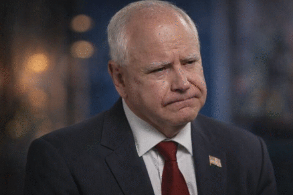 A middle-aged man with short white hair wearing a dark suit and red tie, seated indoors with a serious expression against a softly blurred background.