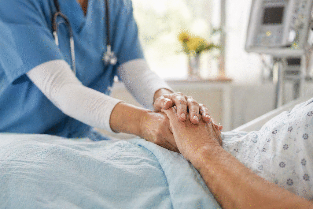 Healthcare professional gently holding an elderly patient’s hand in a hospital bed, with medical equipment softly blurred in the background.