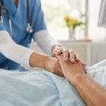 Healthcare professional gently holding an elderly patient’s hand in a hospital bed, with medical equipment softly blurred in the background.
