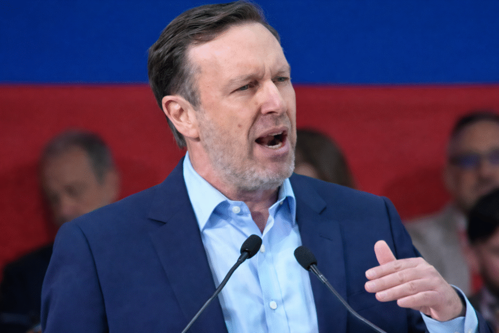Middle-aged man in a navy blazer speaking at a podium with microphones, gesturing with his hand against a blurred red and blue background.